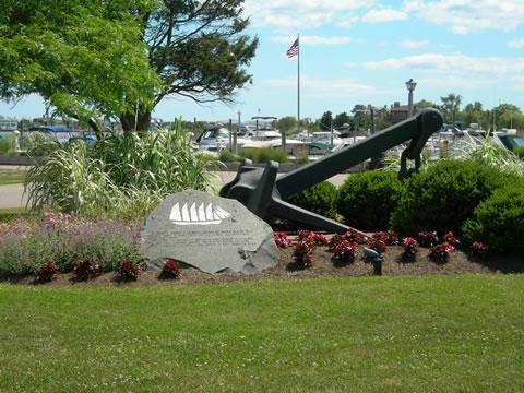 A large ship anchor displayed in a landscaped area with a stone marker, surrounded by flowers and greenery, with boats visible in the background.