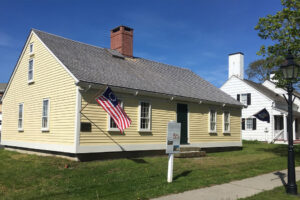 A historic yellow wooden house with a chimney, featuring an American flag hanging from the side, set against a clear blue sky.